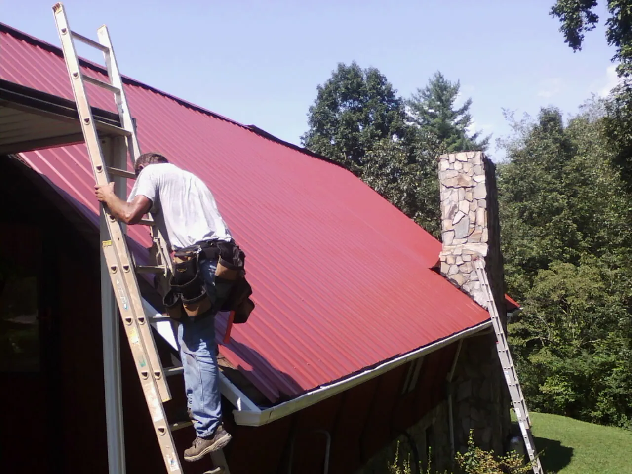 Roofer on a ladder beside a red metal roof and stone chimney — Soffit & Fascia Repair by Roofing Brothers in Ettrick